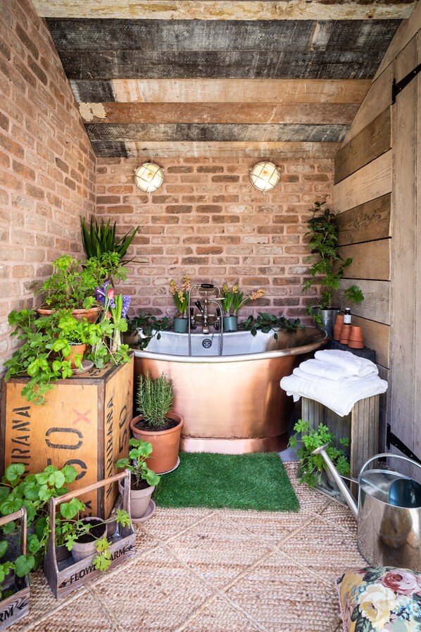 A rustic bathroom with a copper bathtub, a wooden bench, and various potted plants.