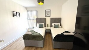 A modern bedroom with two beds, a window with blinds, a ceiling light, and framed pictures on the wall. - Stylish 3BR House 18mins to Peak District/Wifi/Free Parking/Netflix (Ashton-under-Lyne)