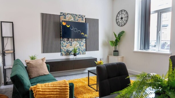 A modern living room with a green sofa, a yellow rug, a black chair, a side table, and a wall-mounted clock. - Stylish 3BR House 18mins to Peak District/Wifi/Free Parking/Netflix (Ashton-under-Lyne)