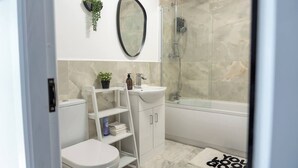 A modern bathroom with a toilet, sink, and shower. There is a shelf with toiletries and a potted plant. - Stylish 3BR House 18mins to Peak District/Wifi/Free Parking/Netflix (Ashton-under-Lyne)
