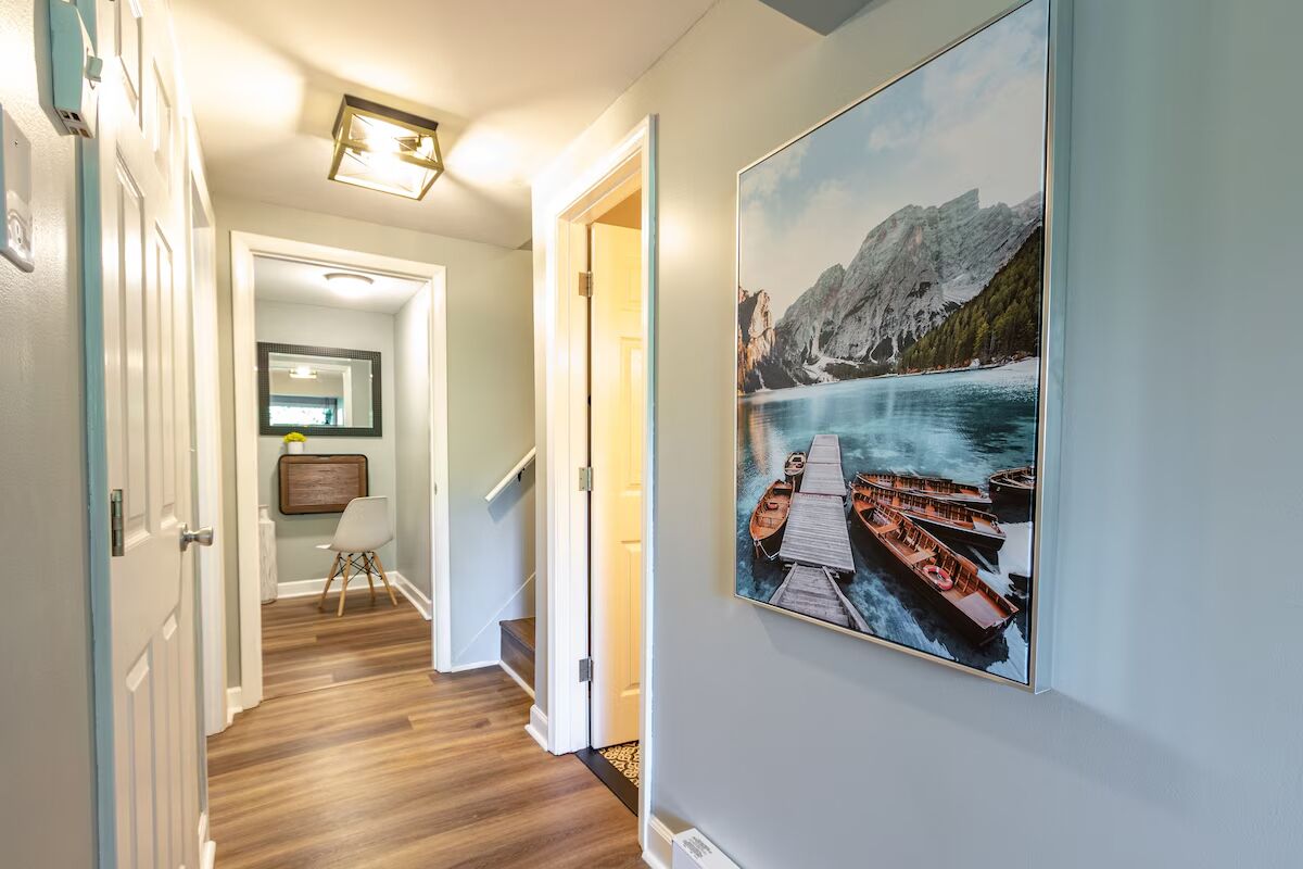 A hallway with wooden flooring, a framed picture of boats on a lake, and a staircase leading to another room.