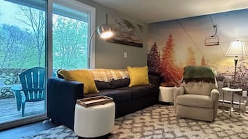 Un salon comprenant un canapé, un fauteuil, un pouf et donnant sur une terrasse par une porte vitrée.