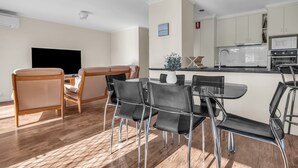Een moderne keuken met witte kasten, een magnetron en een kookplaat. Er is een eethoek met een houten tafel en stoelen, en een televisie in de hoek.