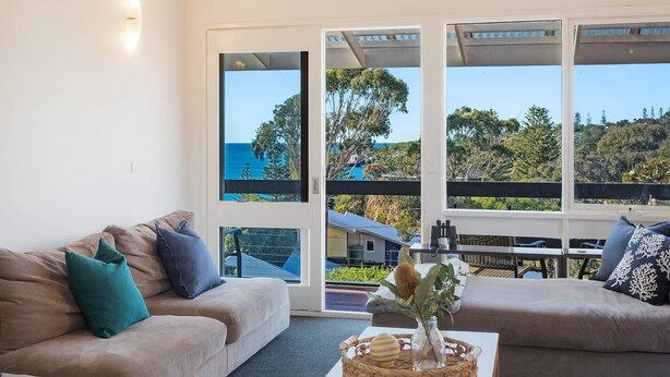 A living room with a beige sofa, a blue cushion, and a coffee table with a woven basket and a vase. Large windows offer a view of a house and trees. - Tathra Beach Shack - Pet Friendly (Tathra)