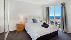 A bedroom with a bed, two bedside tables with lamps, a large window with curtains, and a view of the sea. - Tathra Beach Shack - Pet Friendly (Tathra)