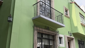 Un edificio de dos pisos con fachada verde, un balcón con barandillas negras y plantas en macetas al frente.