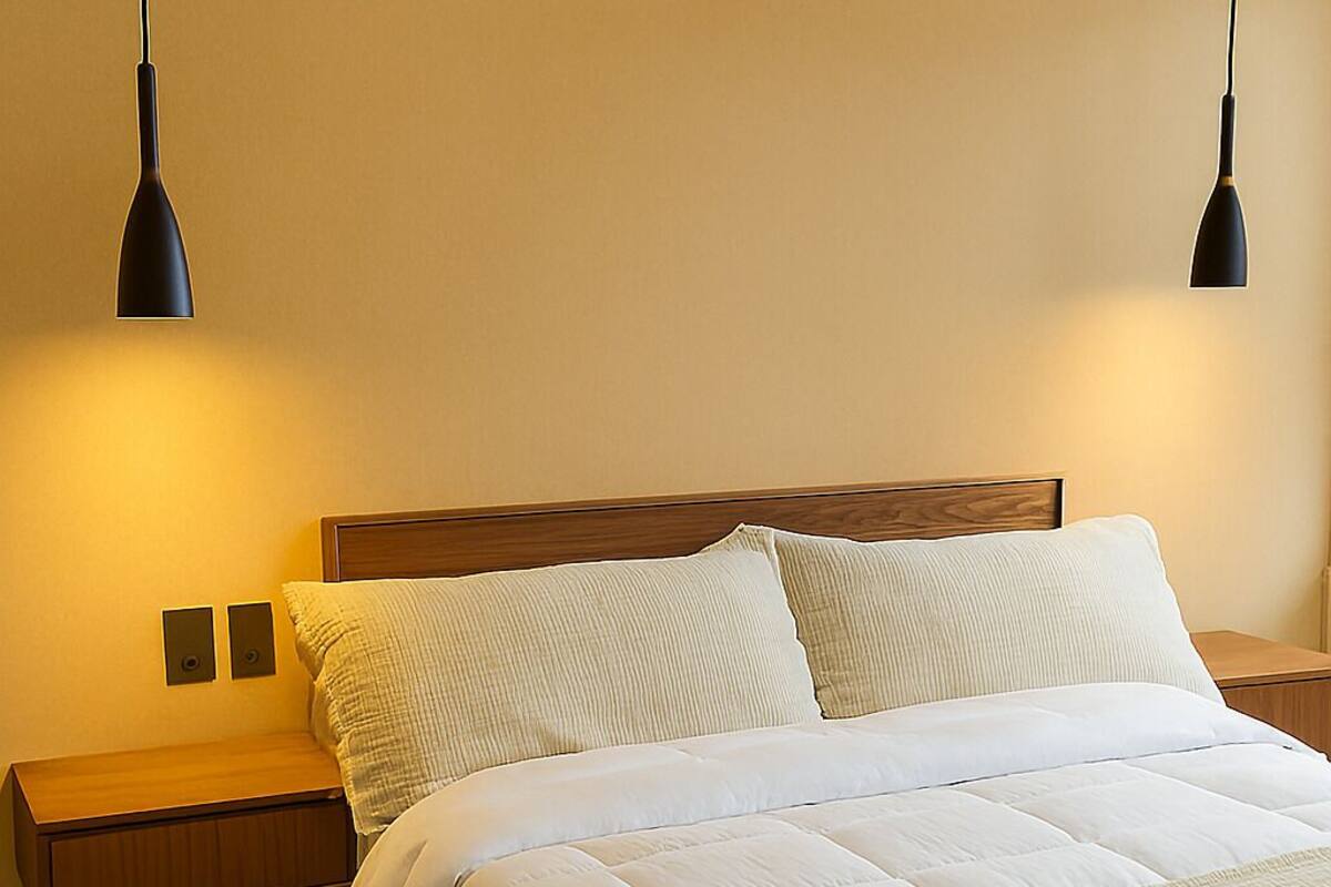 A bed with white bedding, wooden headboard, two bedside tables, and black pendant lights.