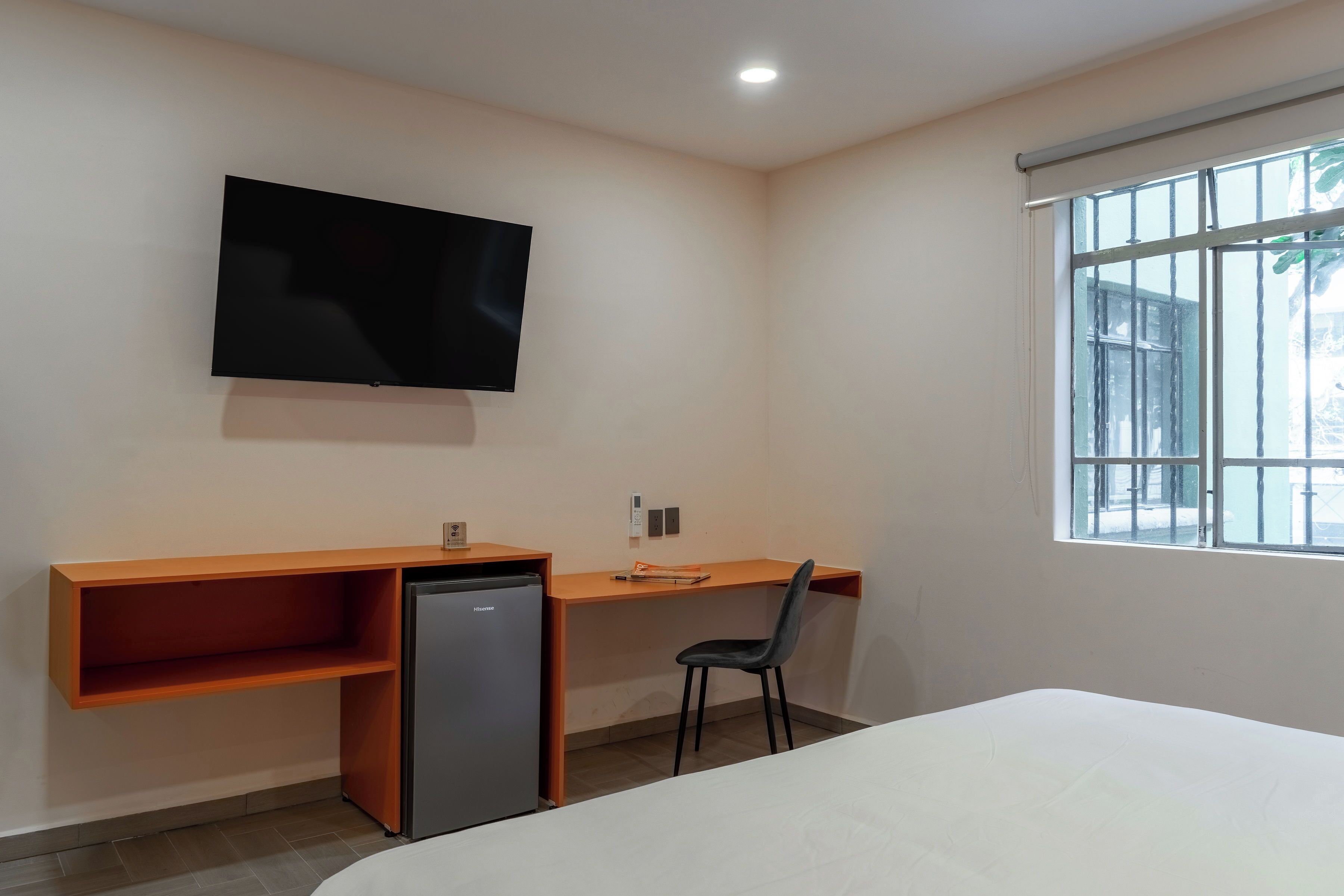 Una habitación con cama, escritorio y silla, televisor montado en la pared y ventana.
