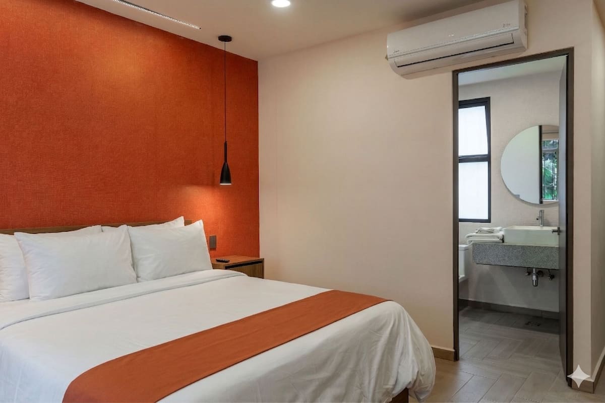 A hotel room with a bed, a bedside table, a hanging light, an air conditioner, and a bathroom visible through an open door.