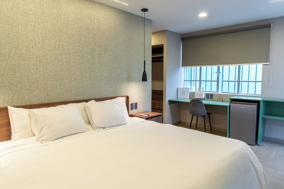 A hotel room with a bed, a desk, a chair, a refrigerator, and a window with blinds.