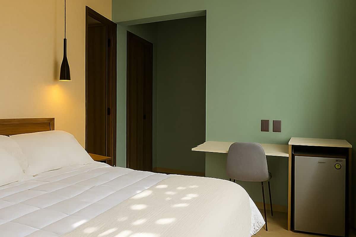 A hotel room with a bed, desk, chair, and a small refrigerator.