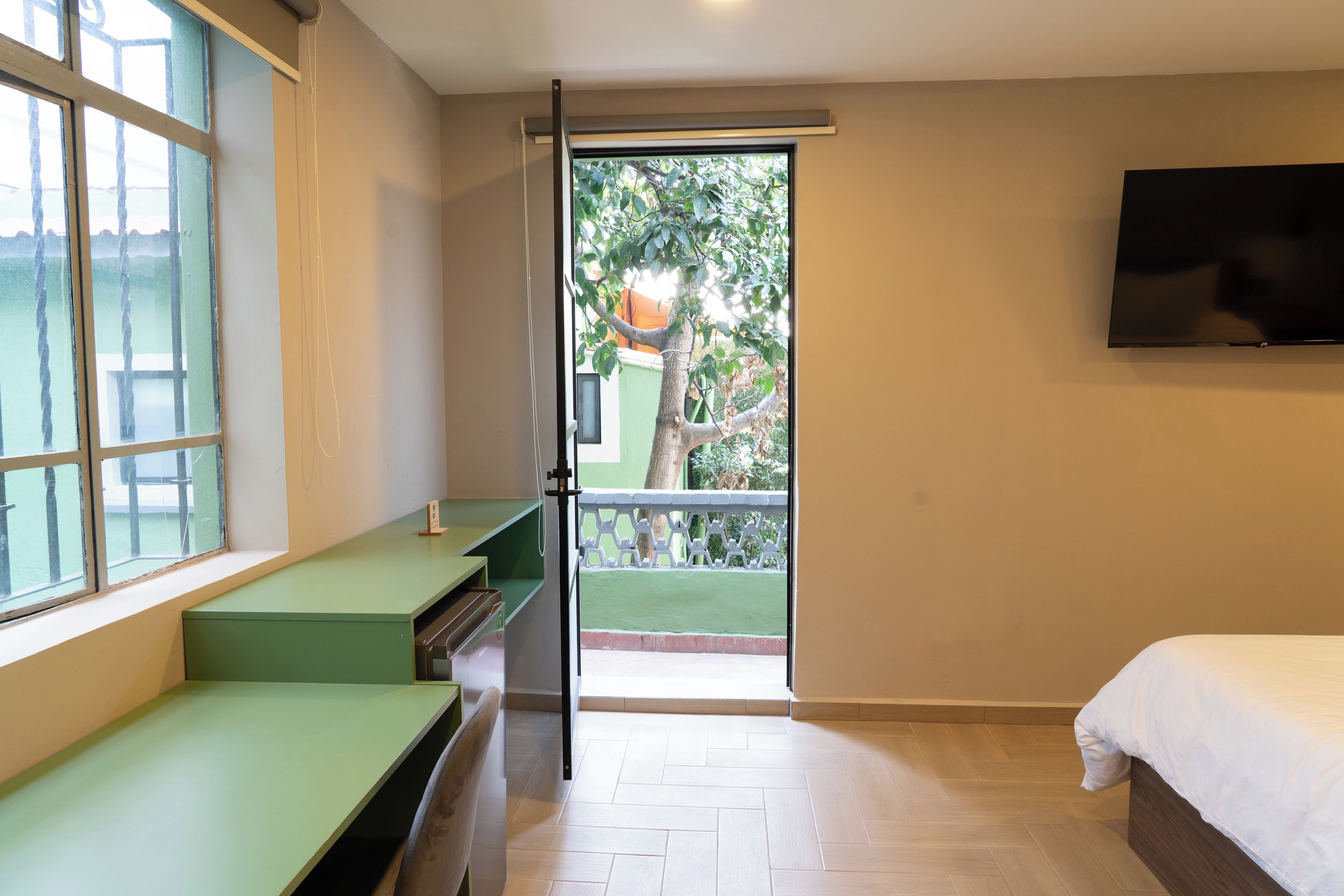 A bedroom with a bed, a TV mounted on the wall, a window with a view of greenery, and a door leading to a balcony.