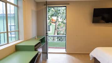 A bedroom with a bed, a TV mounted on the wall, a window with a view of greenery, and a door leading to a balcony.