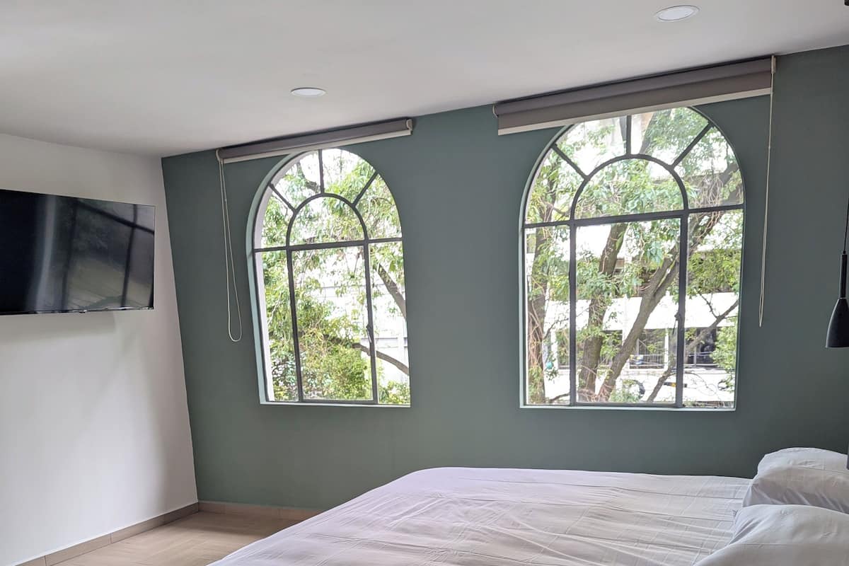 A bedroom with a large bed, two arched windows, a flat-screen TV mounted on the wall, and a dark wooden headboard.