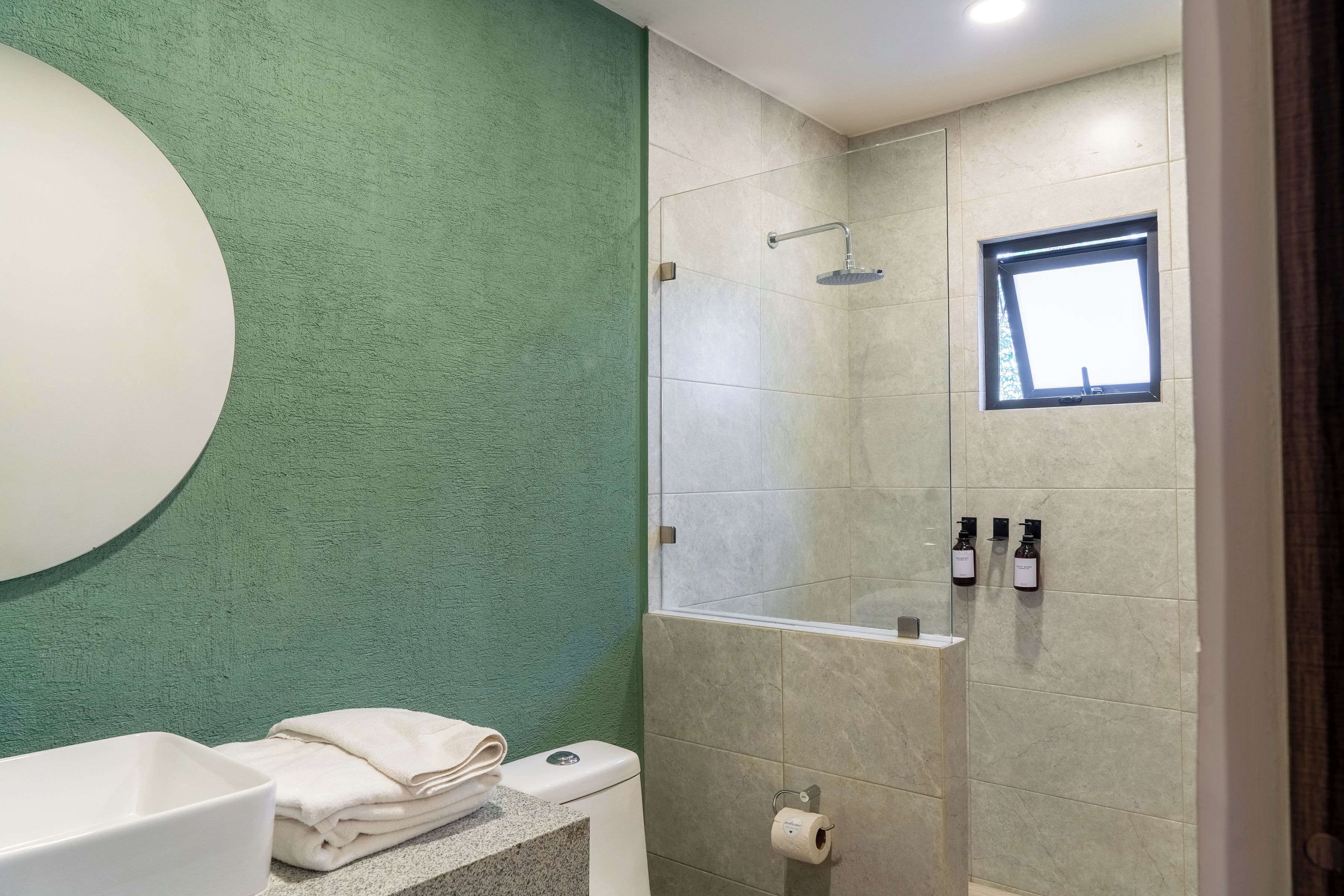 A bathroom with a green wall, a toilet, a sink, a shower, and a window.
