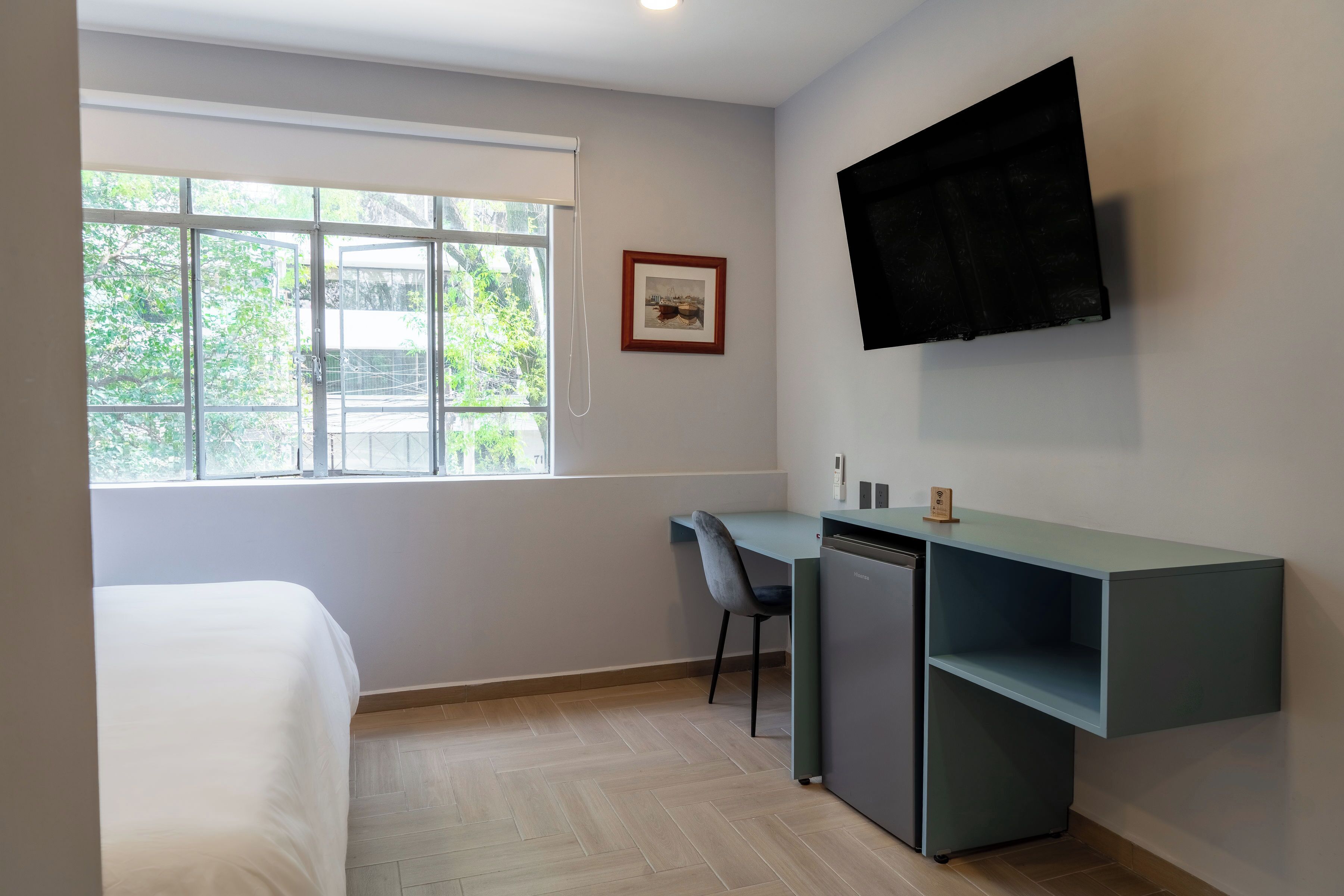 Una habitación con una cama, un escritorio con una silla, un televisor montado en la pared y una ventana con vistas a zonas verdes.