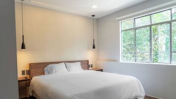 A modern bedroom with a large bed, two bedside tables, a nightstand, a window with a view of greenery, and pendant lighting.