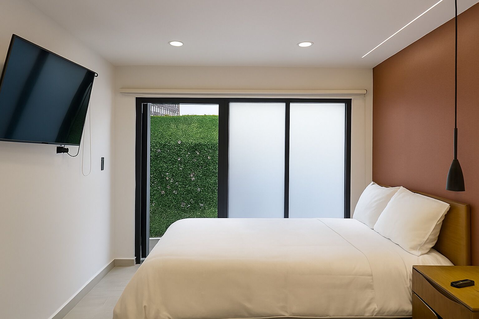 A bedroom with a bed, a TV mounted on the wall, a nightstand, and a view of greenery outside.