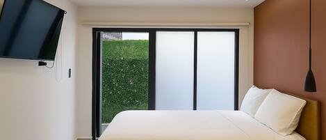 A bedroom with a bed, a TV mounted on the wall, a nightstand, and a view of greenery outside.