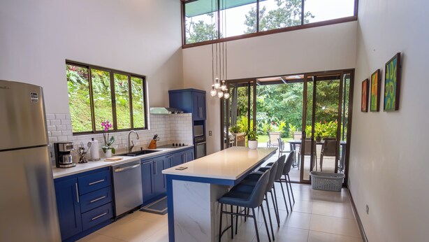 Une cuisine moderne avec des meubles bleus, un îlot en marbre et une grande fenêtre donnant sur un espace verdoyant.