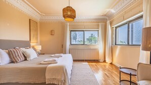 A spacious bedroom with a large bed, a bedside table, a chair, and a window with a view of trees. - 184 - Ocean Views Golf Villa (Cascais)