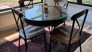 A round glass table with four chairs, a chandelier, and large windows with a view of greenery.