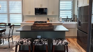 A modern kitchen with light-colored cabinets, a central island with stools, a black refrigerator, and a hanging pendant light.