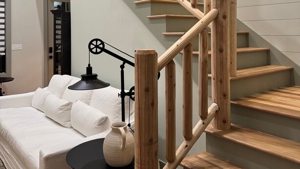 A staircase with wooden railings leading to an upper level, a bed with white bedding, a small table with a lamp, and a wall-mounted light fixture.