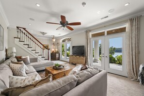 Amplia sala de estar con un sofá grande, una mesa de centro de madera y un televisor. Se ve una escalera que lleva a otro nivel, y ventanas grandes con vistas al exterior.