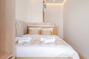 A neatly made bed with white linens and branded towels, a wooden headboard, and a wall-mounted air conditioner.