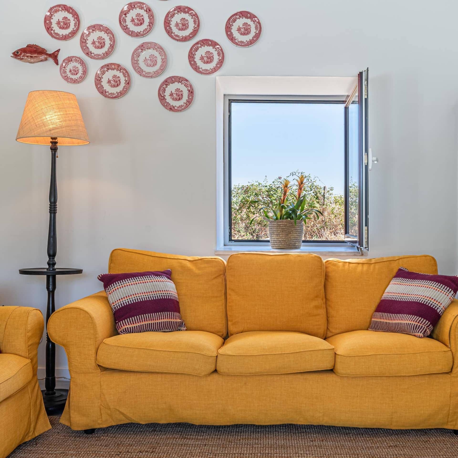 A living room with a yellow sofa, patterned cushions, a floor lamp, decorative plates, and a window with a view of greenery.