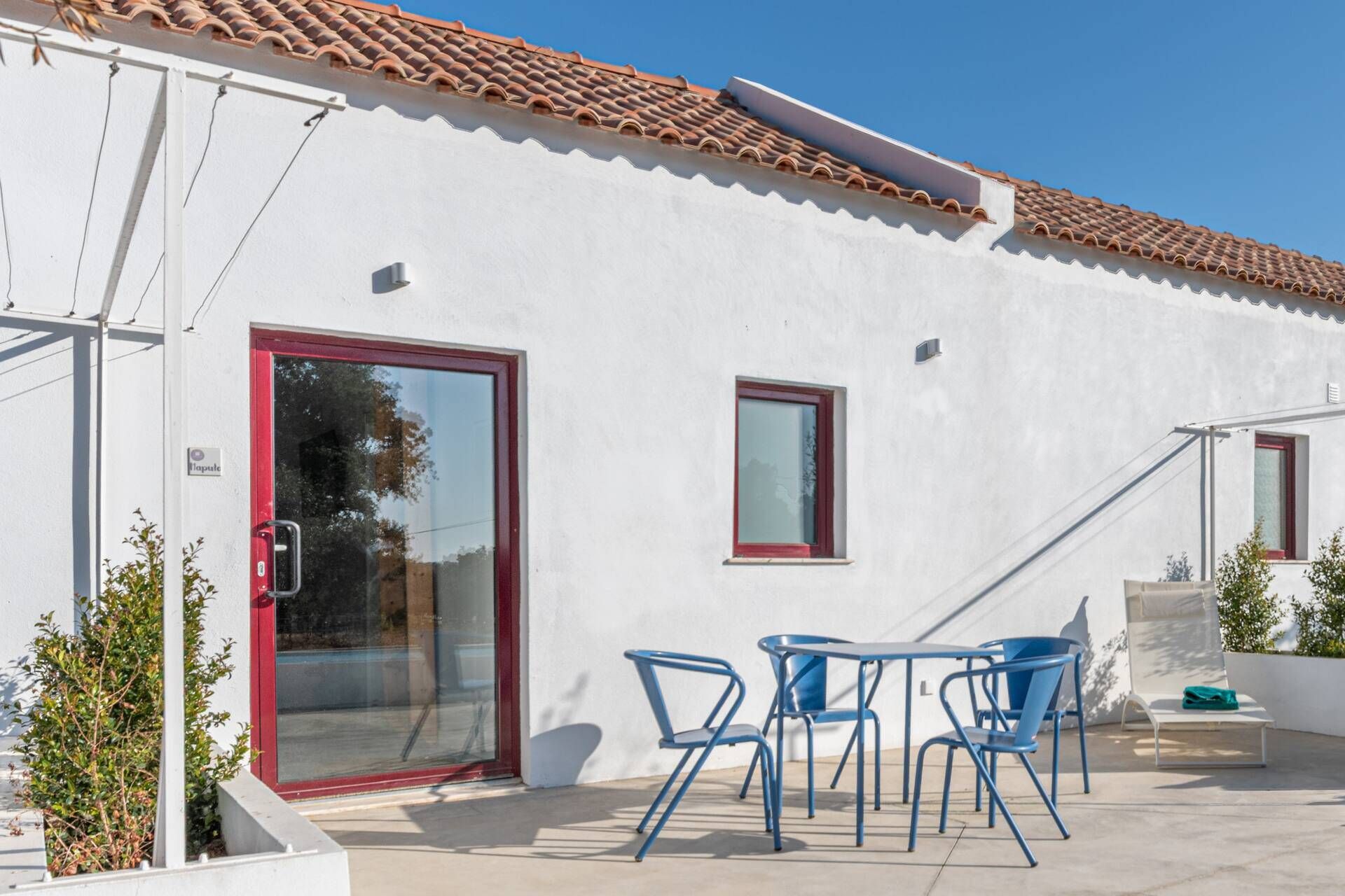 A white building with a red door and windows, a tiled roof, and a patio with blue chairs and a table.