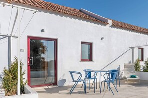 Een wit gebouw met een rode deur en ramen, een pannendak en een terras met blauwe stoelen en een tafel.