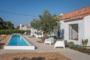 Un espace piscine extérieur moderne, avec une piscine à l’eau d’un bleu limpide, des chaises longues blanches et un bâtiment doté d’un toit en tuiles.