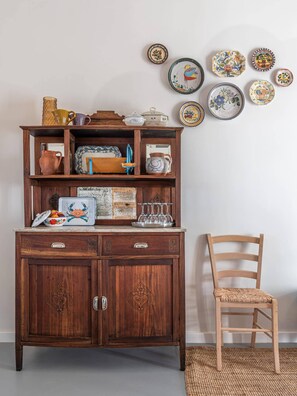 Een houten kast met lades en planken, een rieten stoel en decoratieve borden aan de muur.