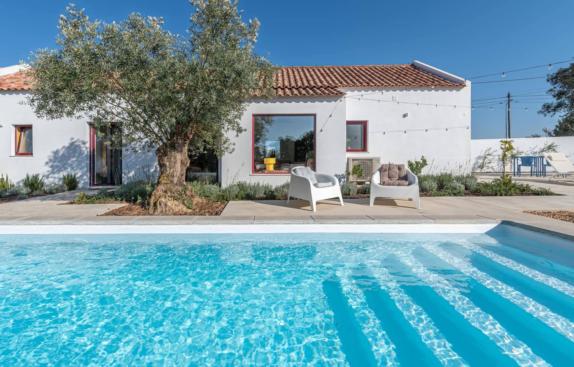Una casa moderna con piscina, dos sillas blancas y un árbol.