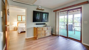 Una habitación amplia con piso de madera, un televisor grande montado sobre un aparador de madera y un ventanal corredizo que da a un espacio exterior con sillas.