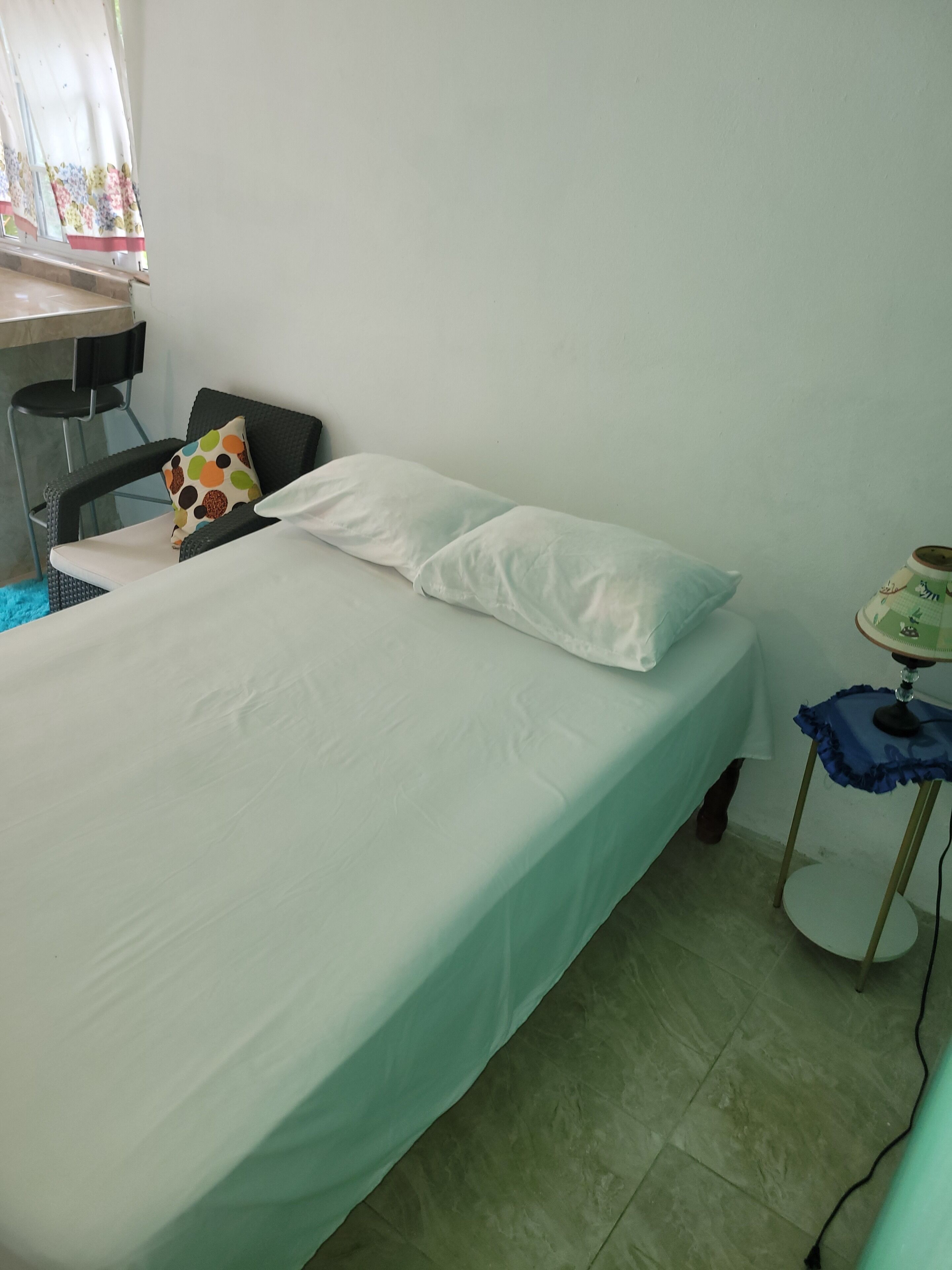 A single bed with a white mattress and pillow, a chair with a polka-dotted cushion, a small table with a green and white patterned top, and a window with floral curtains.
