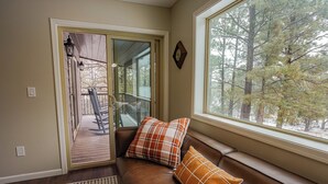 Una habitación acogedora con un sofá de cuero marrón, dos almohadas, un banco de madera afuera y un gran ventanal con vistas a árboles.