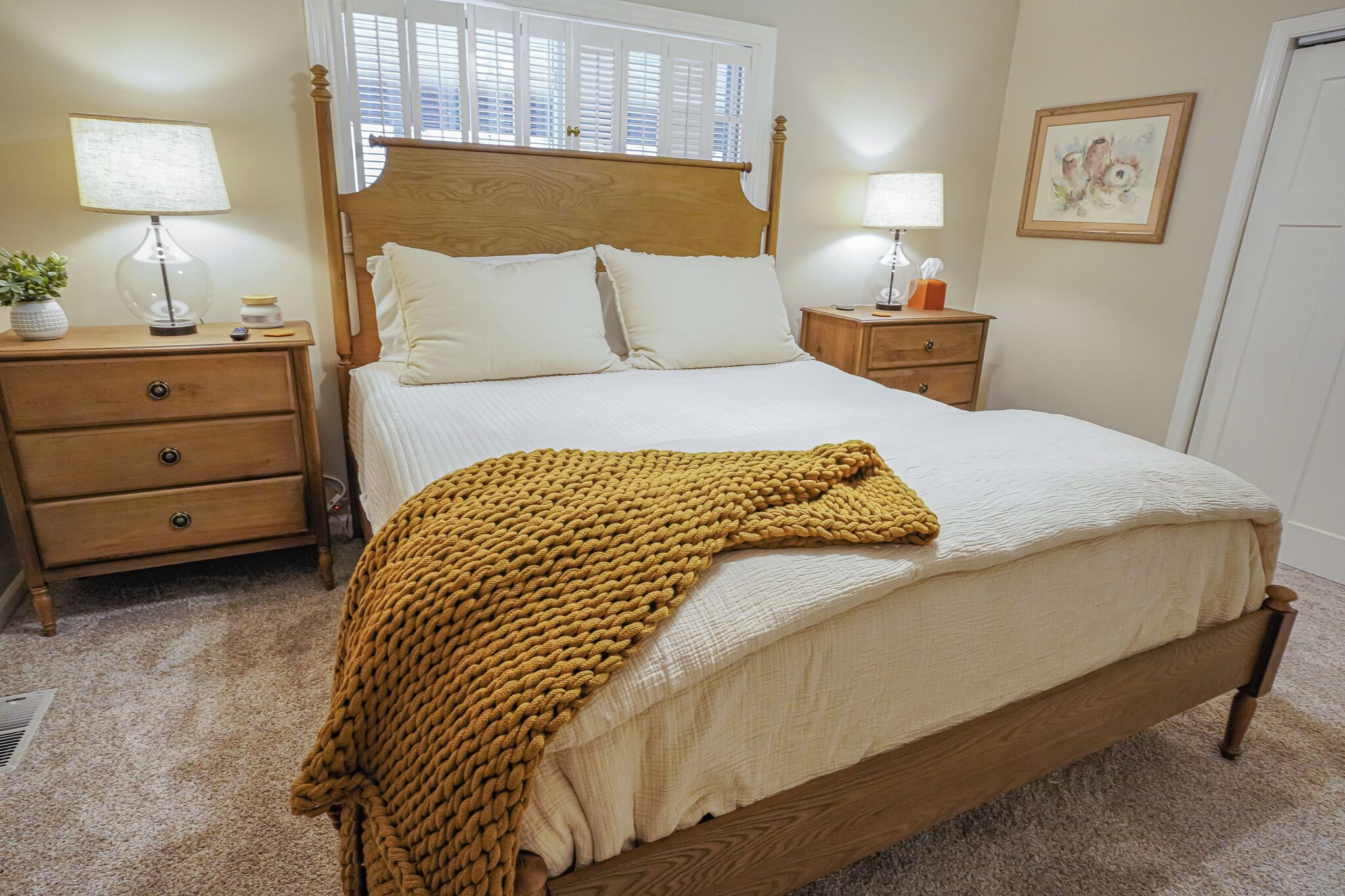 Een netjes opgemaakt slaapkamer met een houten bed, twee nachtkastjes met lampen, een raam met jaloezieën en een lijst met een afbeelding aan de muur.