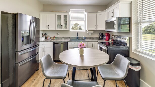 Una cocina moderna con armarios blancos, electrodomésticos de acero inoxidable, una mesa redonda con sillas y una ventana con persianas.