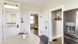 Un comedor con una mesa redonda, sillas y un florero. Se observa una zona de cocina con refrigerador y dispensador de agua.