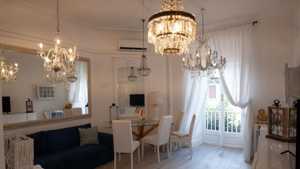 A spacious living room with a dark blue sofa, white chairs, a glass dining table, and multiple chandeliers.