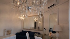 A room with a dark blue sofa, a white side table, a glass coffee table, a white chair, a large mirror, and a chandelier.