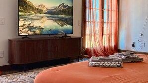 Un dormitorio con una televisión de pantalla grande sobre un soporte de madera, una cama con cubrecama naranja y una ventana con cortinas translúcidas.