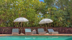 Zona de piscina con sillas de descanso y sombrillas, rodeada de abundante vegetación.