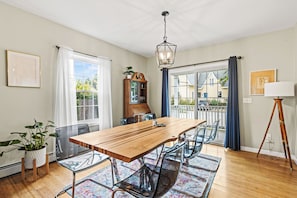 Uma sala de jantar com mesa de madeira, cadeiras transparentes, luminária pendente e vista através de uma porta de vidro.