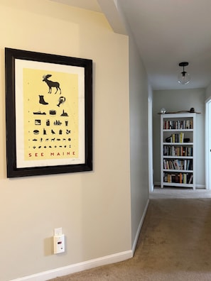 A hallway with a framed picture on the wall, a bookshelf, and a carpeted floor.