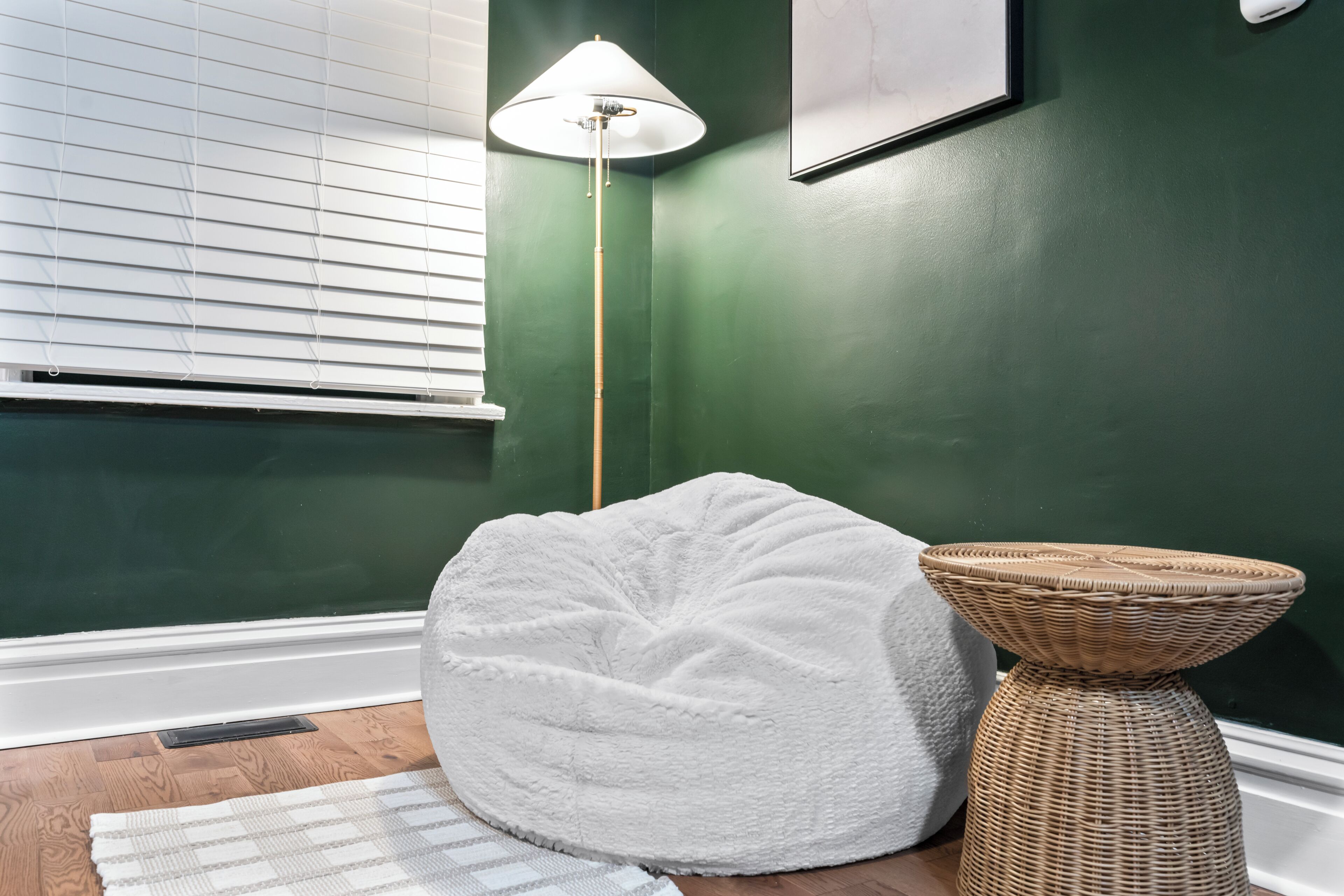 Een kamer met een groene wand, een witte hangzak, een rieten bijzettafeltje en een vloerlamp.