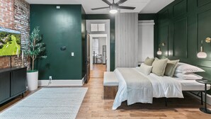 Une chambre moderne avec un grand lit, un ventilateur de plafond, un téléviseur sur le côté gauche et un mur en briques.